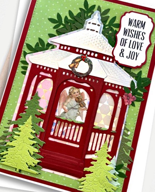 A festive handmade-style greeting card featuring a red gazebo with a violin-playing angel figure inside, green trees, a wreath, and a white label that reads 'Warm Wishes of Love & Joy'.