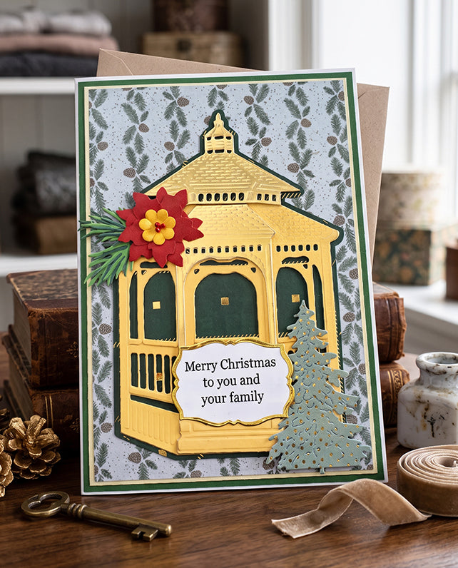 A handmade Christmas greeting card featuring a yellow gazebo with green trim, a red and yellow flower, and a small green tree, with the text 'Merry Christmas to you and your family.' The card is displayed on a wooden surface with vintage props like a key, ribbon, and books.