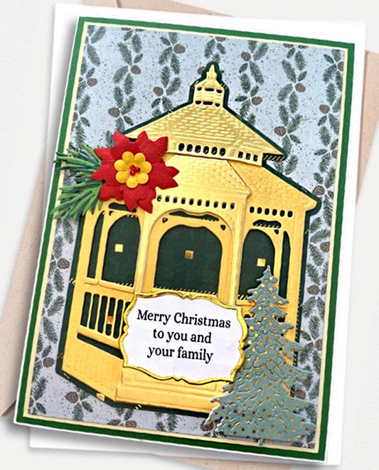 A handmade Christmas greeting card featuring a gold-colored gazebo with a red poinsettia and green pine branch on the left, a blue-green Christmas tree on the right, and a white banner reading 'Merry Christmas to you and your family.' The card has a gray patterned background with green borders and is photographed on a light neutral surface.
