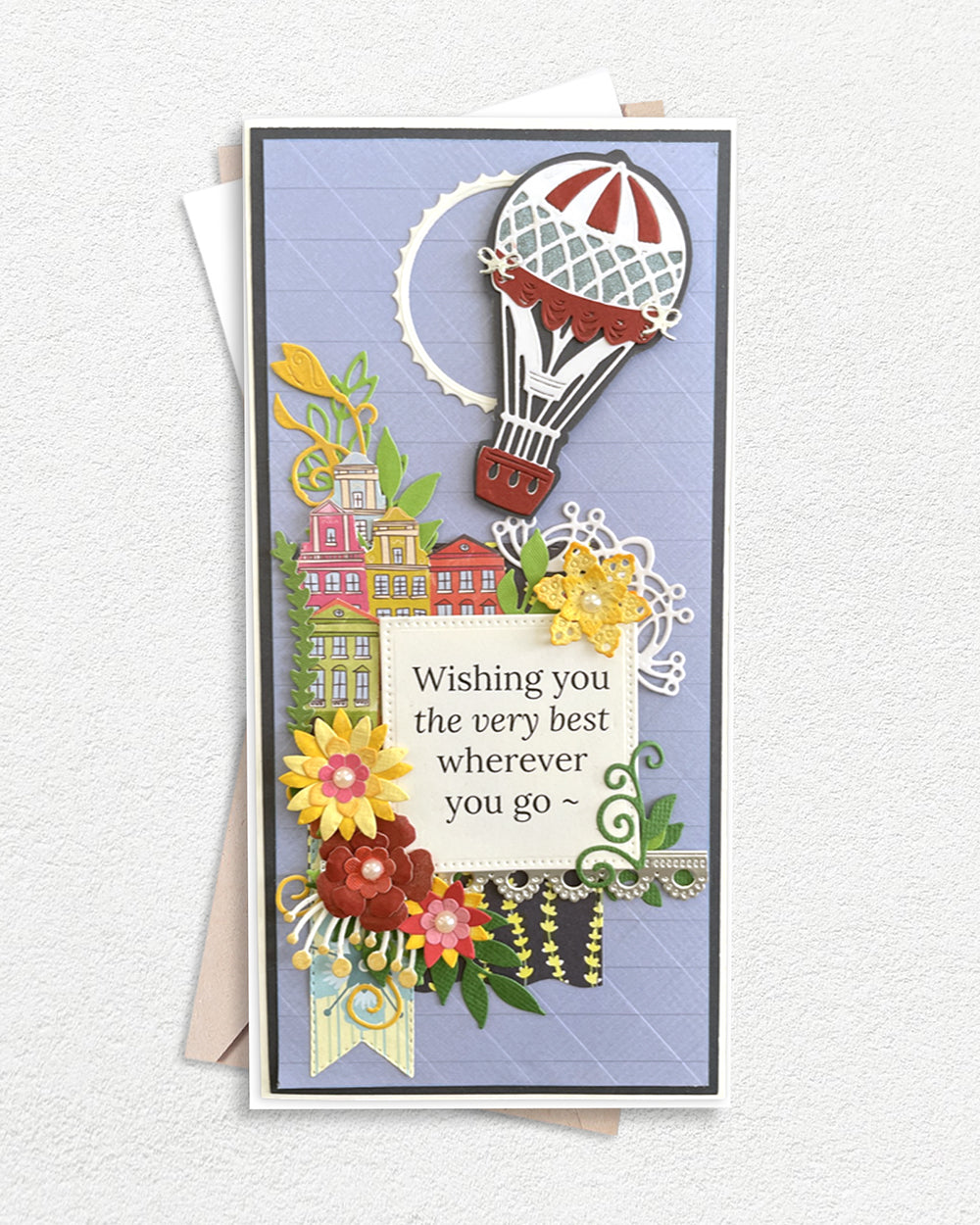 Luxury handmade greeting card featuring a hot air balloon above a colourful village scene, surrounded by layered die-cut flowers in red, yellow and green, on a soft blue quilted background, with a sentiment panel reading "Wishing you the very best wherever you go".