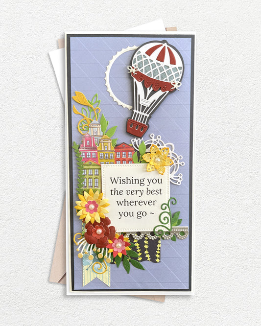 Luxury handmade greeting card featuring a hot air balloon above a colourful village scene, surrounded by layered die-cut flowers in red, yellow and green, on a soft blue quilted background, with a sentiment panel reading "Wishing you the very best wherever you go".