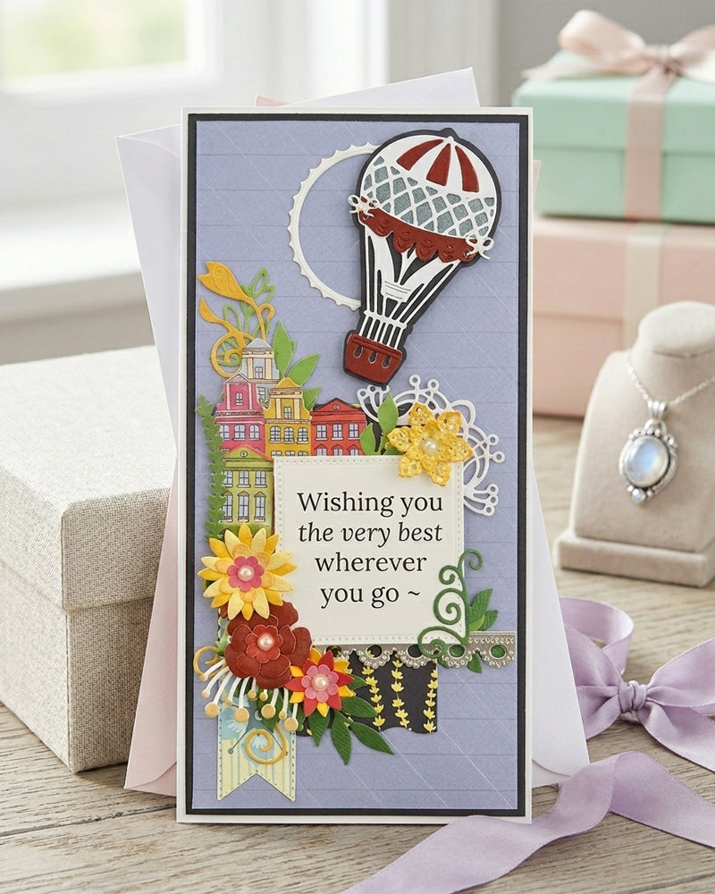 Handmade luxury greeting card displayed upright beside a wrapped gift with ribbon, showing full card design with hot air balloon and village scene, made in New Zealand by Del Cards