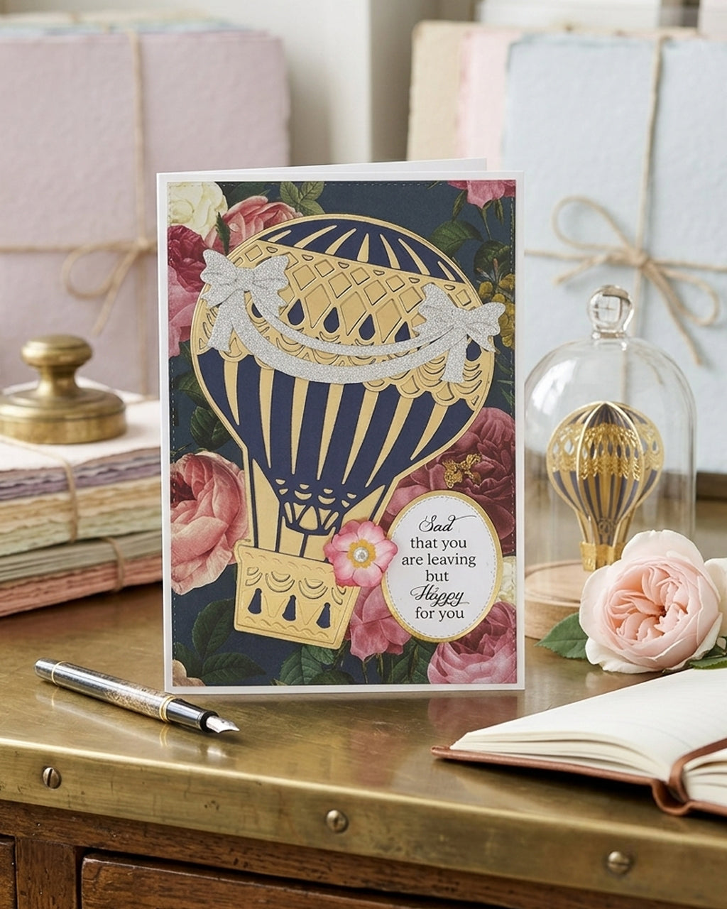 A square greeting card featuring a navy blue and gold hot air balloon illustration with pink roses and a white message bubble reading 'Sad that you are leaving but hope for you.' Made with love by Del Cards NZ.