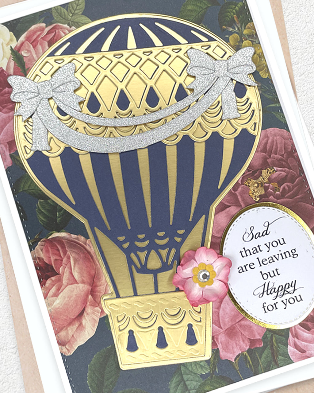 A close-up of a decorative greeting card featuring a large gold and navy hot air balloon over pink roses, with a small pink flower and a round white tag that reads, 'Sad that you are leaving but Happy for you.'.