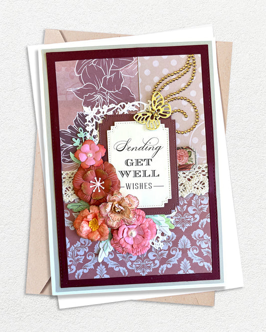 Handmade luxury get well card on a deep burgundy cardstock base layered with a soft mauve and white large-scale floral stamped background paper. A gold swirl flourish trails across the upper portion, adding elegance and movement. Hand-crafted paper flowers in deep rose, coral, peach and burnt orange cluster at the lower centre. 
