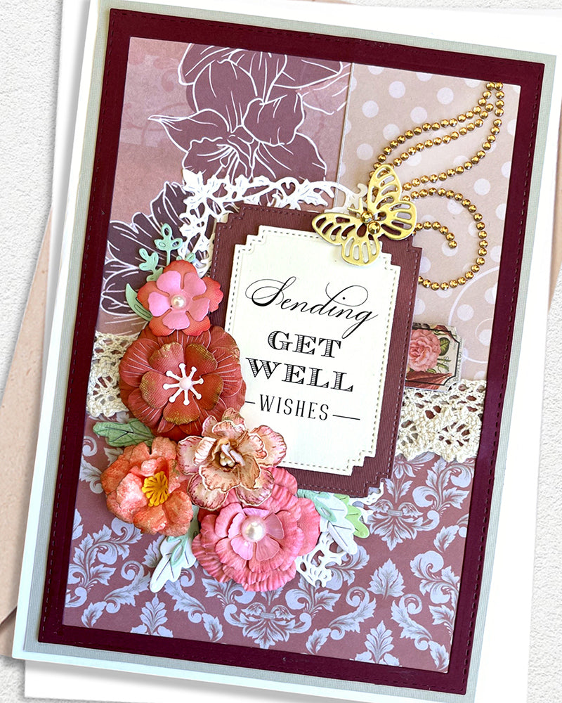 Close-up of a handmade luxury get well card showing clustered paper flowers in deep rose, coral and peach tones with a gold swirl flourish above and a white sentiment frame reading "Sending Get Well wishes." Mauve floral stamped background and white lace die-cut border visible. Handmade greeting card made in New Zealand.