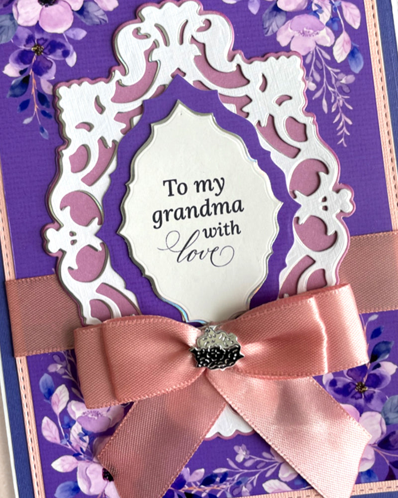 Luxury handmade card for grandmother featuring deep purple layered cardstock, white ornate die-cut frame, hand-applied purple paper florals, and a wide satin bow in dusty rose pink. Sentiment reads "To my grandma with love." Premium handmade greeting card, New Zealand.

