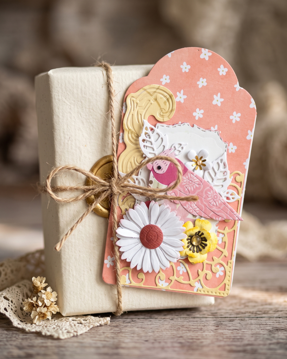A styled gift tag attached to a cream-colored book or box with twine, featuring layered paper designs in peach, white, pink, yellow, and gold on a rustic wooden surface.