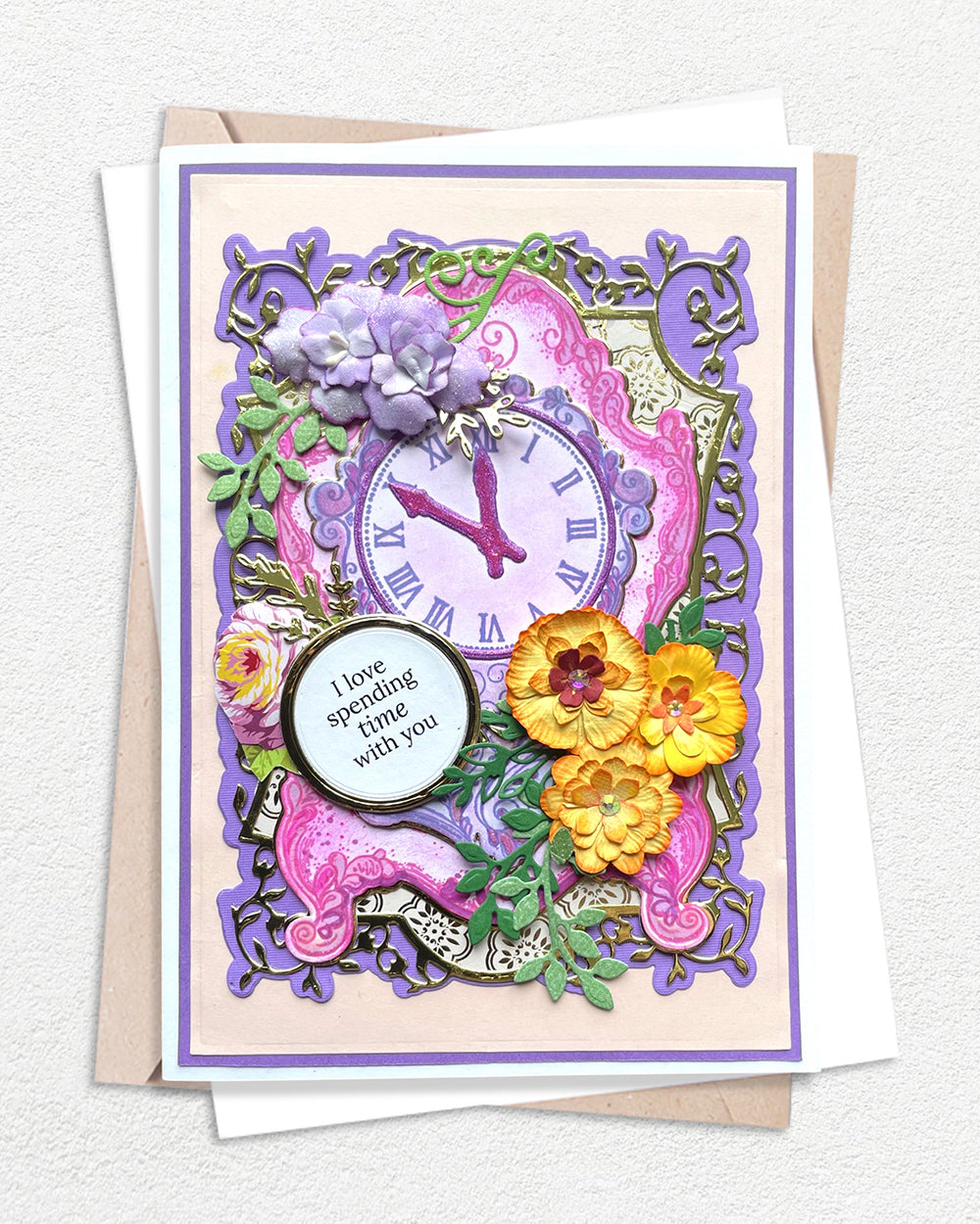 Luxury handmade card featuring an ornate purple clock face surrounded by layered die-cut flowers in yellow, pink and purple, with intricate green foliage, set within a decorative purple frame on a pink and cream background, with a circular sentiment panel reading "I love spending time with you"
