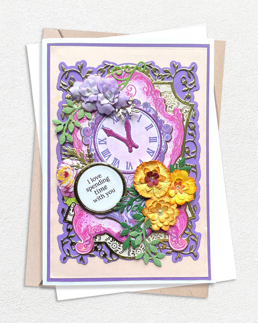 Luxury handmade card featuring an ornate purple clock face surrounded by layered die-cut flowers in yellow, pink and purple, with intricate green foliage, set within a decorative purple frame on a pink and cream background, with a circular sentiment panel reading "I love spending time with you"