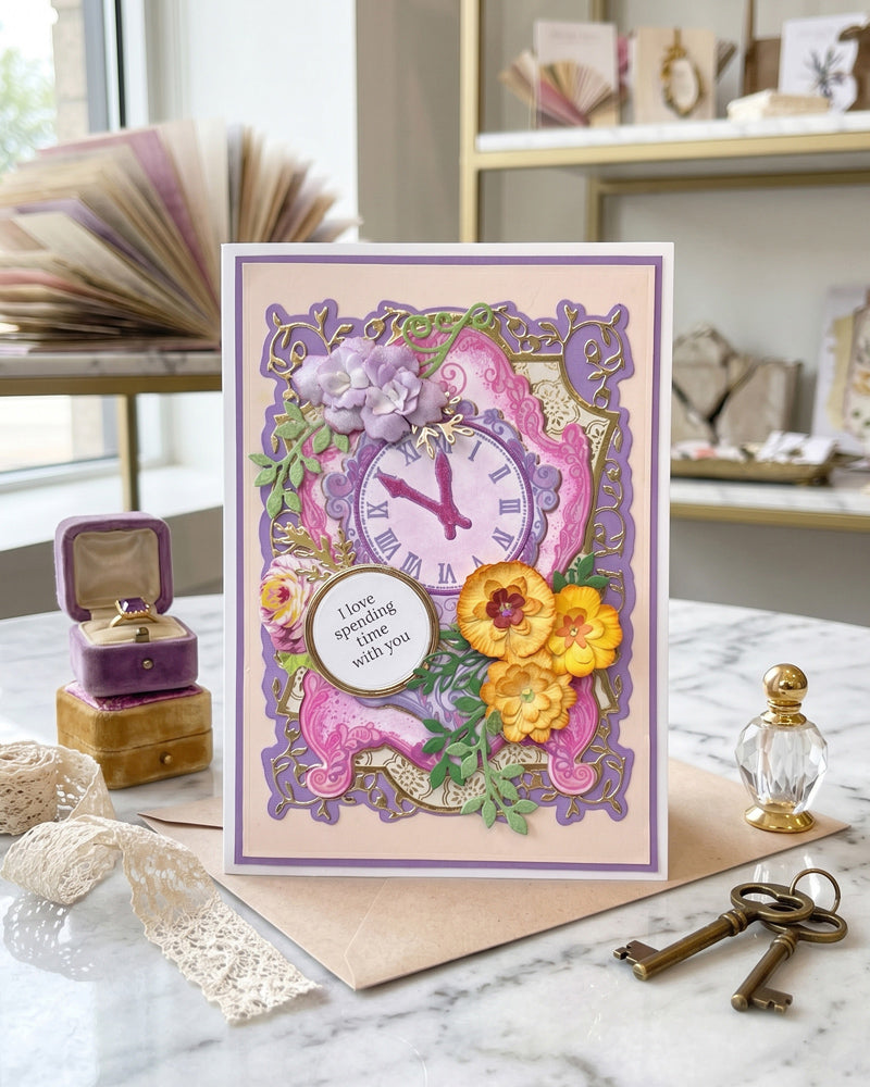 Handmade luxury card displayed upright on a surface beside vintage keys and soft fabric, showing full card design with purple clock, layered flowers and ornate frame made by Del Cards NZ.