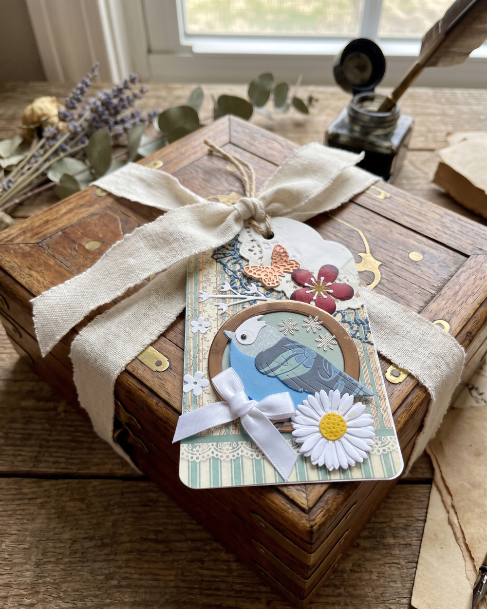 A decorative handmade gift tag featuring a blue bird, white daisy, and butterfly embellishments, tied with a cream ribbon to a rustic wooden box on a wooden table.