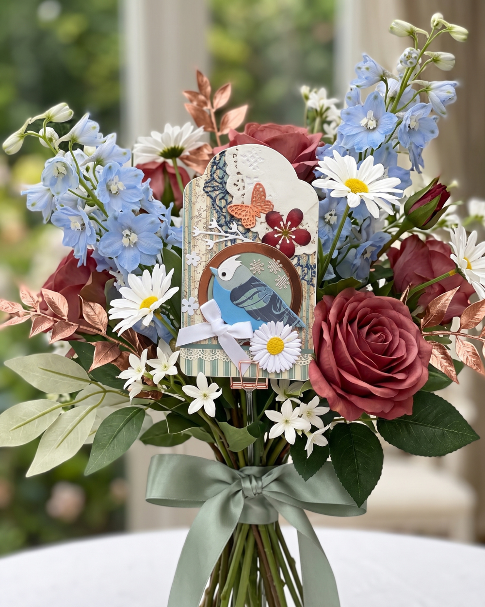 A decorative floral arrangement in a vase with a pale sage ribbon, featuring blue delphiniums, white daisies, burgundy roses, green leaves, and a central card with a blue bird and butterfly motif.