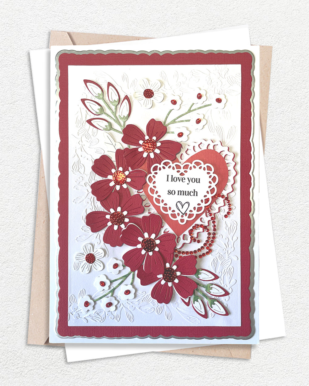 Luxury handmade love card featuring large dimensional deep red flowers with jewel centres, delicate red foliage sprays with crystal embellishments, and a lace-edged heart sentiment panel reading "I love you so much" on a white embossed background with red border.