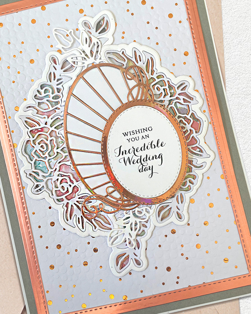 A close-up of a handmade greeting card featuring a white oval center with the text 'Wishing You an Incredible Wedding day' in black script, surrounded by layered white floral and leaf cutouts with a metallic rose-gold center frame. The card has a peach border, a gray inner border, and scattered gold foil dots on a white background.