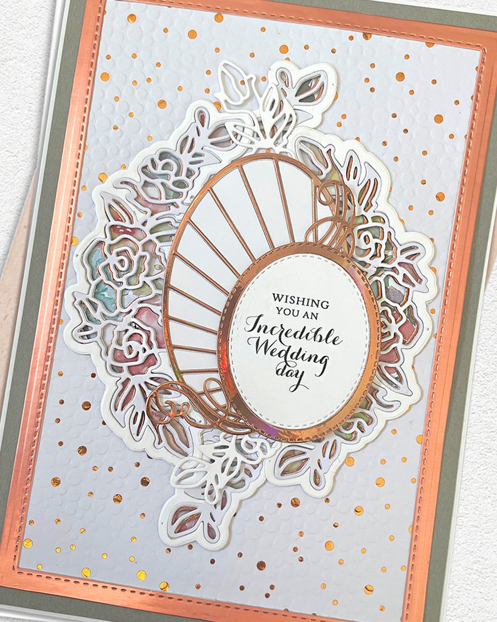 A handmade wedding greeting card featuring a layered die-cut floral design with a central oval frame and the printed message 'WISHING YOU AN Incredible Wedding day.' The card has a white base with scattered gold and pink confetti, a peach border, and a gray outer edge, photographed against a light textured background.