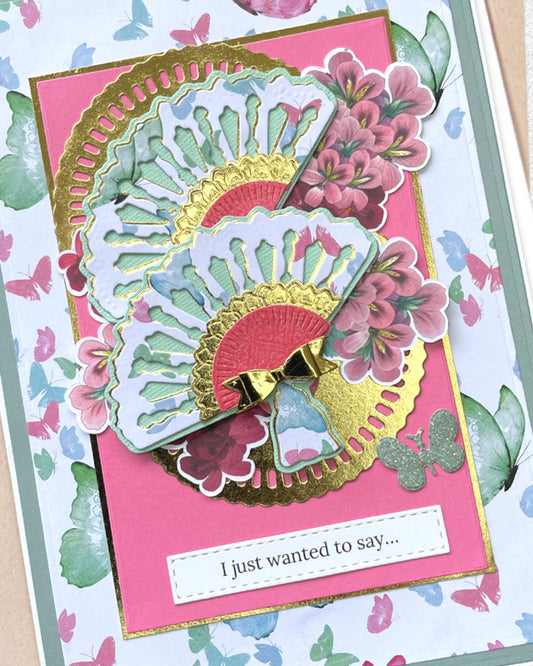 Close-up of a handmade luxury card showing the upper portion of a large layered paper fan in mint, white and gold with deep pink floral accents, set against a hot pink panel and white butterfly-patterned background. Handmade greeting card made in New Zealand.