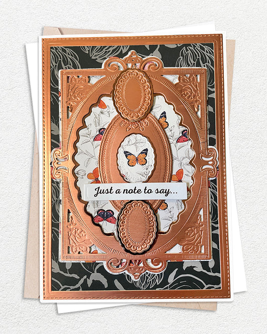 Handmade luxury greeting card with a dramatic dark floral background in deep charcoal, layered with an ornate copper-toned embossed die-cut frame featuring scrollwork and oval window details. A smaller oval at the top holds a delicate hand-applied paper butterfly. The centre panel reveals a white floral paper layer beneath the copper frame, with scattered mini butterfly accents. A ribbon-style sentiment banner reads "Just a note to say..." in fine script. White envelope visible beneath.
