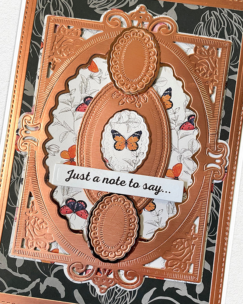 Full front view of a handmade copper and charcoal greeting card featuring an intricate embossed copper die-cut frame, dark floral background, white floral underlayer, scattered butterfly details and a "Just a note to say..." sentiment banner. A sophisticated, versatile handmade card made in New Zealand — available for sale, perfect for thinking-of-you, thank you or everyday occasions.