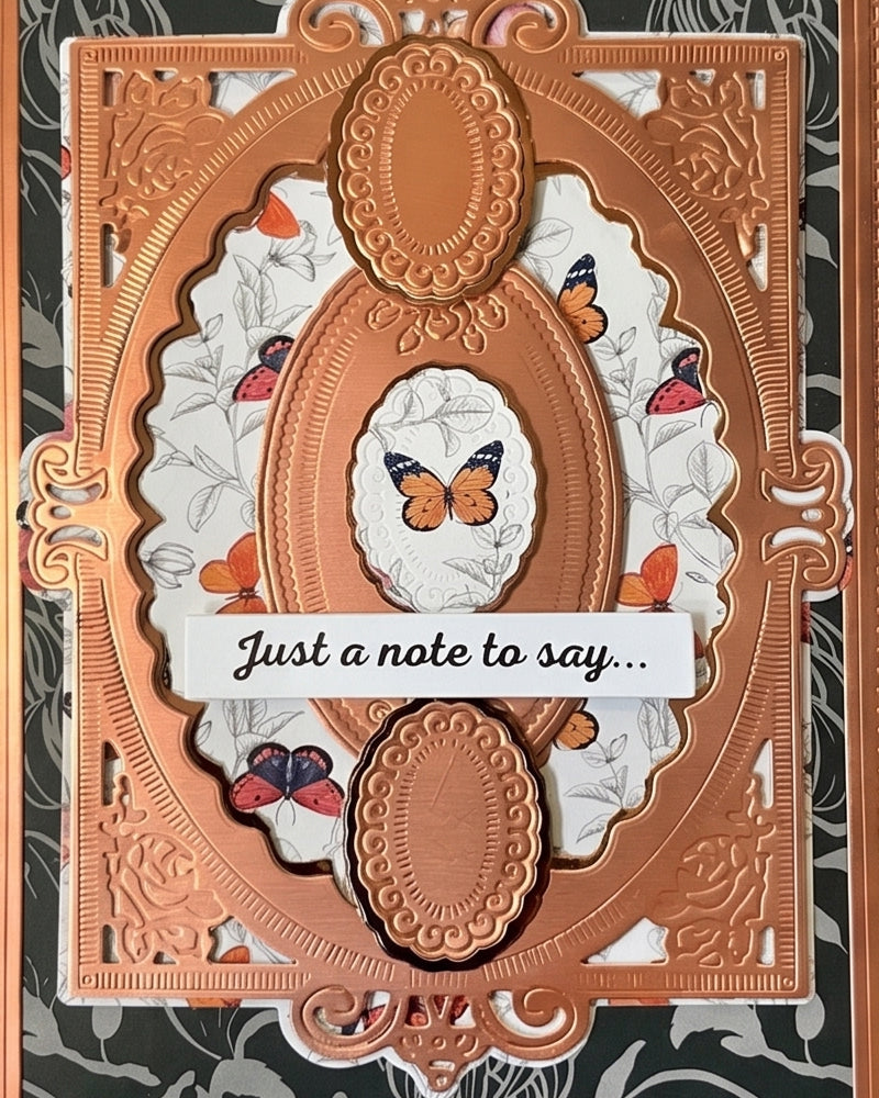Close-up of a handmade luxury card showing the upper portion of an ornate copper embossed die-cut frame with a small oval window featuring a delicate paper butterfly, set against a dark charcoal floral background with white floral underlayer detail. Handmade greeting card made in New Zealand.