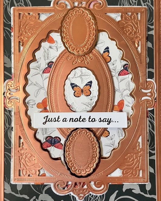 Close-up of a handmade luxury card showing the upper portion of an ornate copper embossed die-cut frame with a small oval window featuring a delicate paper butterfly, set against a dark charcoal floral background with white floral underlayer detail. Handmade greeting card made in New Zealand.