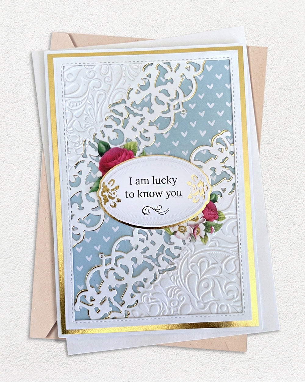 Handmade luxury friendship card on a soft powder blue cardstock base with a gold foil mat. An intricate large-scale white lace and heart filigree die-cut overlay covers the full card face, creating rich texture and depth. A gold-edged oval centrepiece holds a cream sentiment panel reading "I am lucky to know you" in elegant script, with a small hand-applied red and pink rose spray as a delicate accent. White and kraft envelopes visible beneath. Handmade luxury friendship card made in New Zealand.