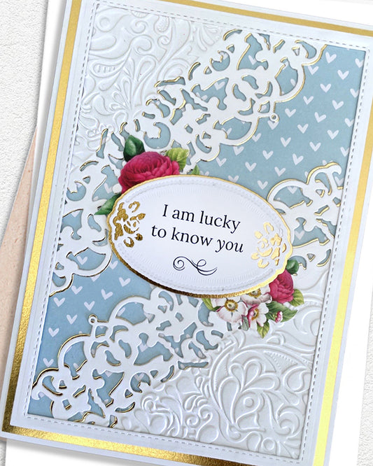 Close-up of a handmade luxury friendship card showing a gold-edged oval sentiment panel reading "I am lucky to know you" with a small red and pink rose spray accent, set against an intricate white lace and heart filigree die-cut overlay on a powder blue background. Handmade greeting card made in New Zealand.

