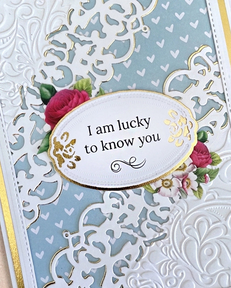 Full front view of a handmade powder blue and white friendship card featuring a full-coverage white lace heart filigree die-cut overlay, gold foil mat, and a gold-edged oval sentiment reading "I am lucky to know you" with a delicate rose accent. An elegant, refined handmade friendship card made in New Zealand by Del.