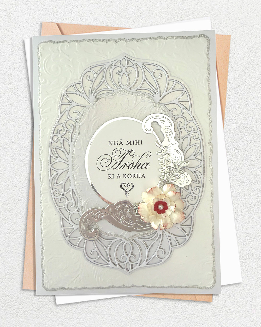 Handmade greeting card with ornate silver filigree frame and dimensional paper flowers, reading "Ngā Mihi Aroha ki a Kōrua" in Te Reo Māori, shown with cream and peach envelopes