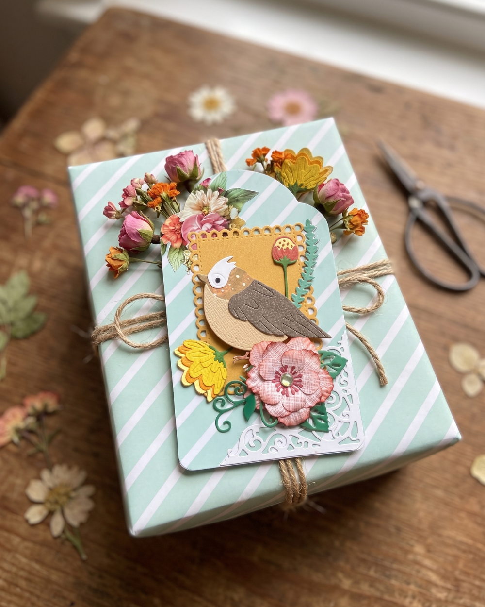 A wrapped gift box with mint green and white diagonal stripes, decorated with a layered paper card featuring a bird and pink flowers, tied with twine on a rustic wooden table.