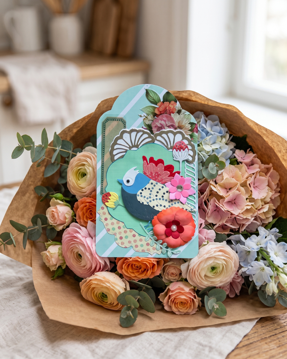 A decorative gift tag featuring a colorful peacock illustration and floral details, placed on top of a wrapped bouquet of pink, peach, orange, and blue flowers with greenery, set in a softly lit indoor setting.