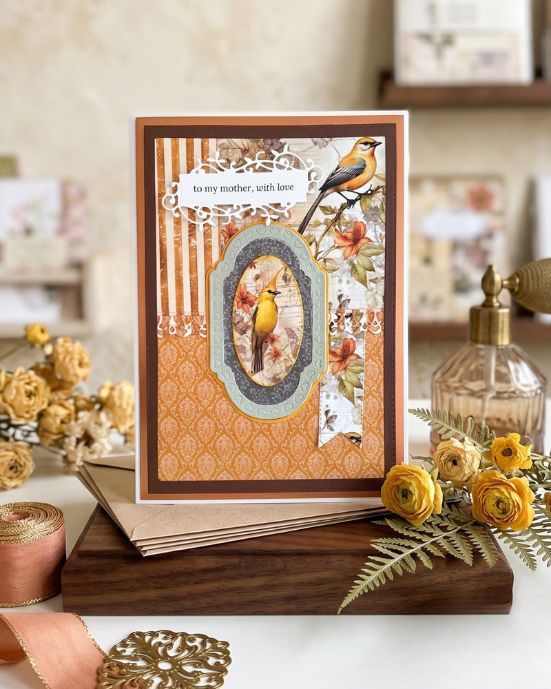 Handmade luxury mother's card styled in a warm home setting, propped against a wooden surface with natural dried flowers alongside. The amber and chocolate tones of the card complement the warm, organic styling. Handmade in Auckland, New Zealand.

