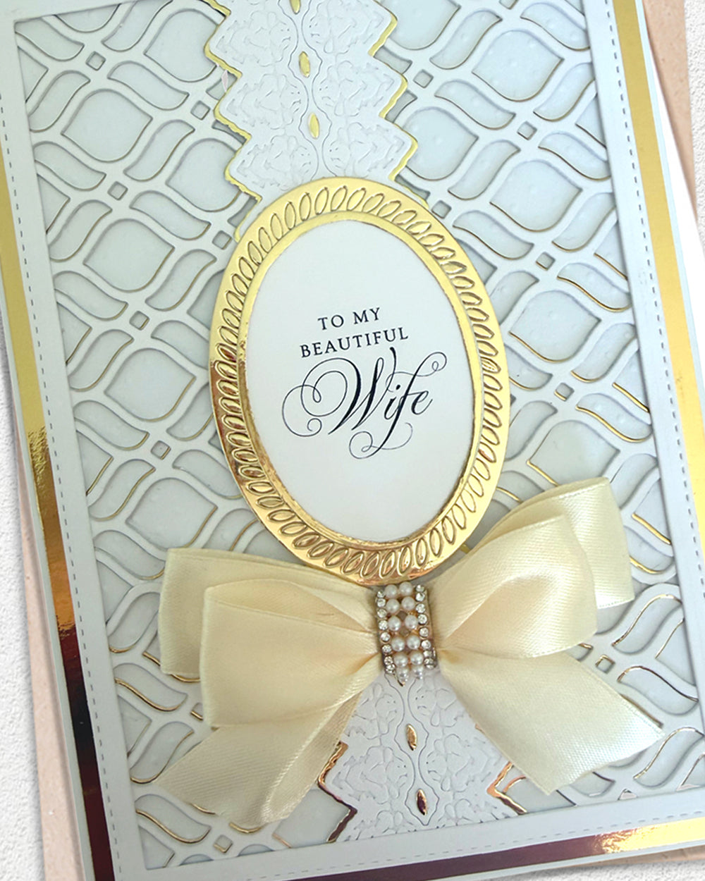 A close-up of a decorative greeting card or paper craft featuring a white embossed geometric pattern, a gold oval border, the phrase 'TO MY BEAUTIFUL Wife' in black script, and a cream satin bow with a rhinestone center.