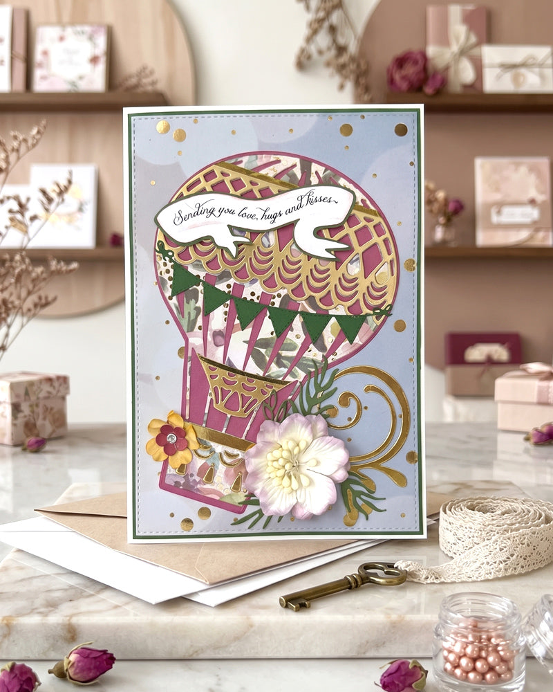 Handmade luxury card displayed upright on a warm styled surface beside a decorative teacup and scattered petals, showing full hot air balloon card design in deep pink and gold, made in NZ by Del Cards