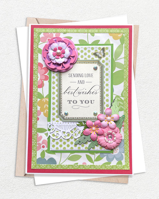 Handmade luxury greeting card on a deep pink cardstock base layered with a bright green botanical floral patterned paper featuring illustrated leaves and flowers. A cream sentiment frame at the centre reads "Sending Love and best wishes to you" in mixed serif and script lettering. A large layered deep pink glitter paper flower sits upper left, with clusters of smaller pink cherry blossom-style flowers and green leaves spilling across the lower right. 