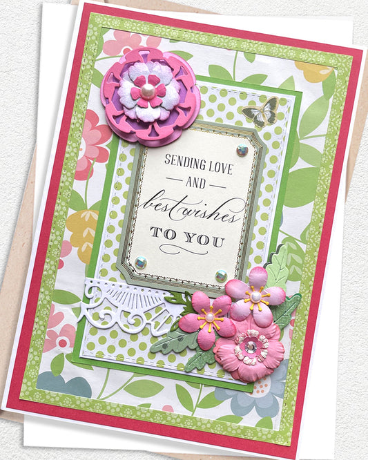 Close-up of a handmade luxury card showing a large layered deep pink glitter paper flower in the upper left, a cream sentiment frame reading "Sending Love and best wishes to you" and clusters of smaller pink flowers with green leaves. Bright green botanical patterned background. Handmade greeting card made in New Zealand by Del Cards.