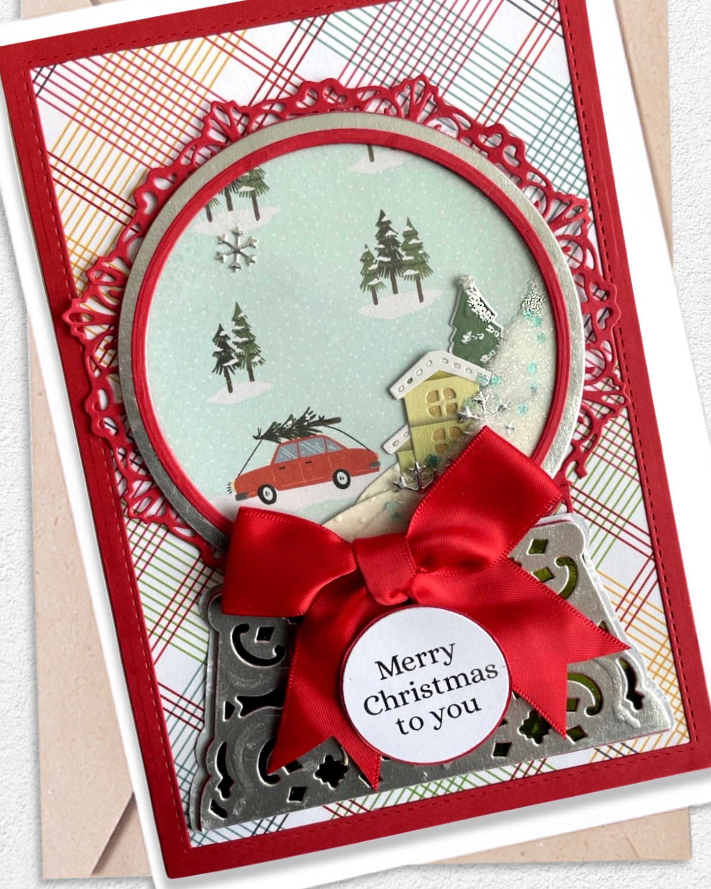 A handmade Christmas greeting card featuring a red ribbon bow, a circular window with a winter village scene, and a small round tag that reads 'Merry Christmas to you'.