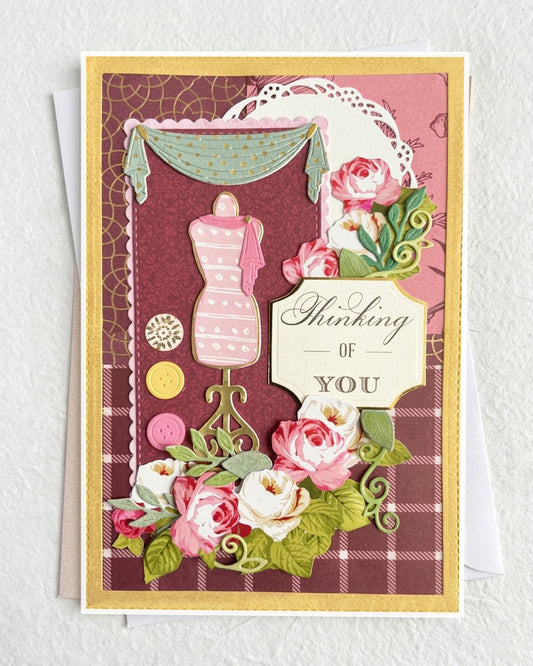 Handmade luxury thinking-of-you card on a gold cardstock base layered with a deep burgundy plaid patterned paper. A central pink dressmaker's mannequin on an ornate stand takes centre stage, framed by a teal curtain drape and white doily detail at the top. Scattered gold and yellow button embellishments add a playful craft-inspired touch. Lush hand-applied pink and white paper roses with green leaves spill across the lower portion.