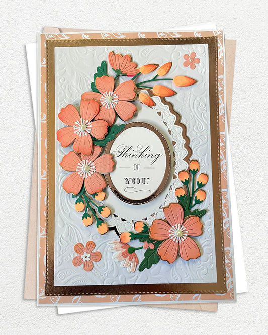 Handmade luxury thinking-of-you card on a white hand-embossed cardstock base with a warm gold cardstock mat. A abundant wreath of hand-crafted coral and peach five-petal paper flowers with orange bud accents and deep green leaves surrounds a white scalloped oval frame. The oval centrepiece holds a sentiment reading "Thinking of You" in layered serif and bold lettering. Card shown in clear protective sleeve with kraft and white envelopes beneath. Made by Del Cards from New Zealand.