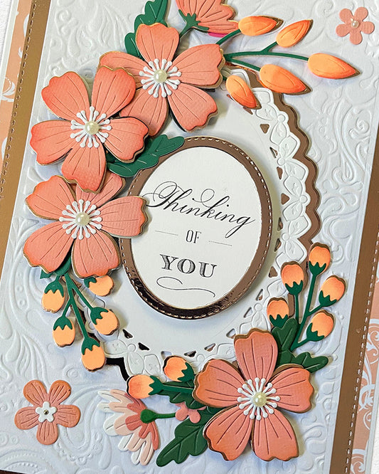 Close-up of a handmade luxury card showing the upper half of a lush coral and peach paper flower wreath with orange bud accents and green leaves, surrounding a white scalloped oval with "Thinking of YOU" sentiment. Hand-embossed white cardstock background visible. Handmade greeting card made in New Zealand.

