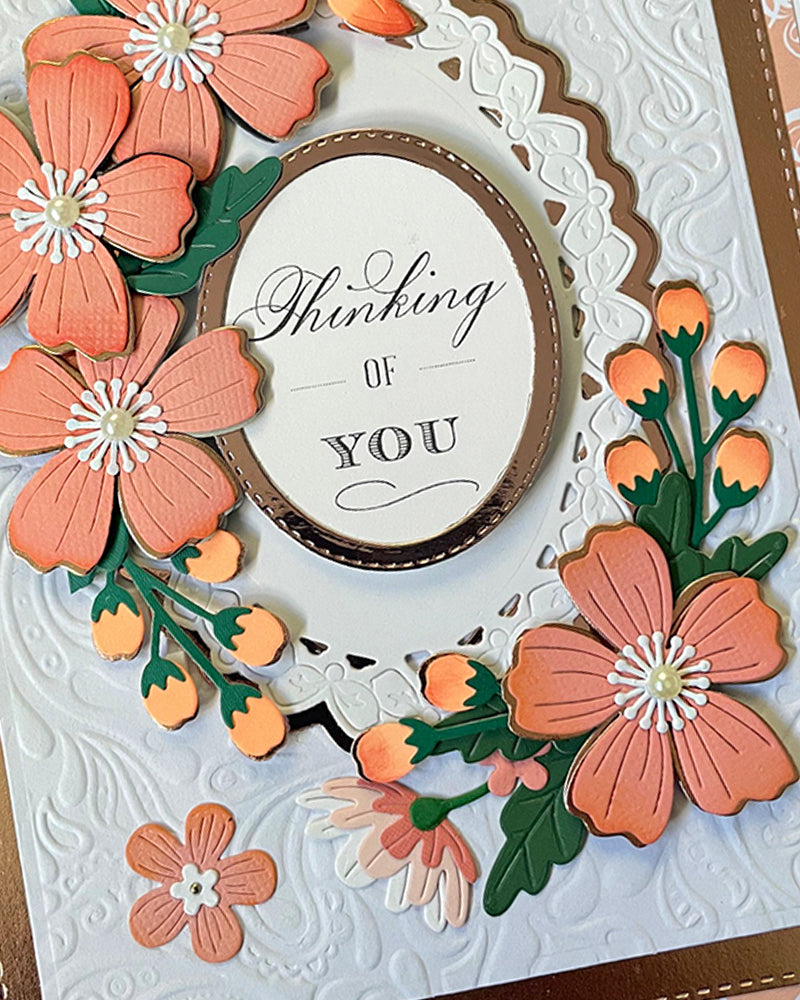 Full front view of a handmade white and coral greeting card featuring a full wreath of hand-crafted peach paper flowers, deep green leaves, orange bud details and a scalloped oval "Thinking of You" sentiment centrepiece on a hand-embossed background. A beautiful handmade thinking-of-you card made in NZ by Del Cards.