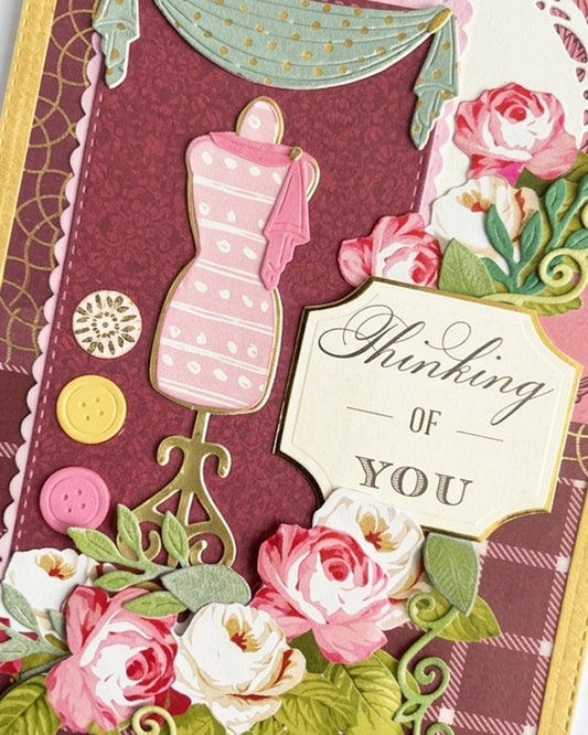 Close-up of a handmade luxury card showing a pink dressmaker's mannequin on an ornate stand, teal curtain drape, white doily detail, scattered button embellishments and a cream sentiment tag reading "Thinking of You." Pink and white paper roses with green leaves visible below. Handmade greeting card made in New Zealand by Del.