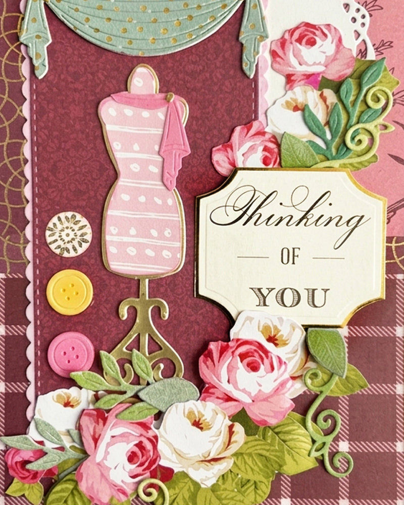 Full front view of a handmade burgundy and gold thinking-of-you card featuring a plaid background, central pink dressmaker's mannequin, teal curtain detail, button accents, lush pink and white paper roses and a "Thinking OF YOU" sentiment tag. A whimsical, feminine handmade card made in New Zealand  by Del Cards NZ.