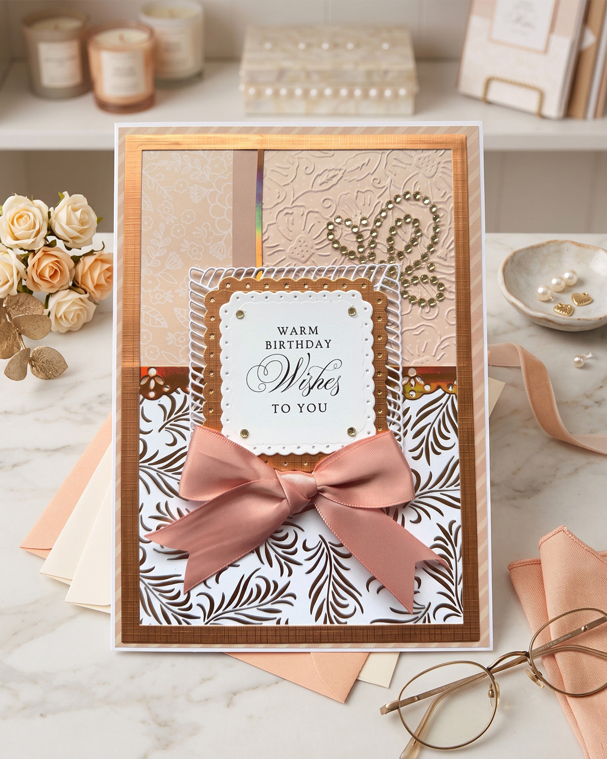 A handmade square greeting card with layered cream, blush pink, and white patterns, a large dusty pink satin bow, and a central white label reading 'Warm Birthday Wishes to You.' The card is styled on a marble surface with blush ribbons, flowers, candles, and eyeglasses in the background.
