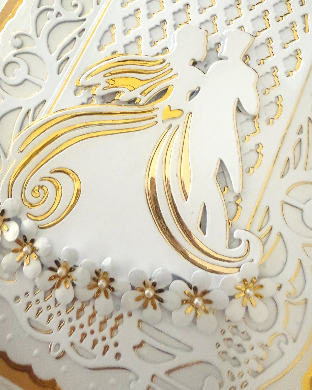 A close-up of a white and gold decorative card or paper craft featuring a stylized bride and groom silhouette, floral cutouts, and ornate lace-like patterns.