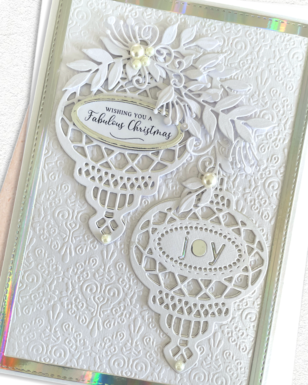 A close-up of a handmade Christmas greeting card featuring intricate white layered die-cut ornaments, floral elements, and small pearl accents on a textured white cardstock background with a holographic shimmering border.