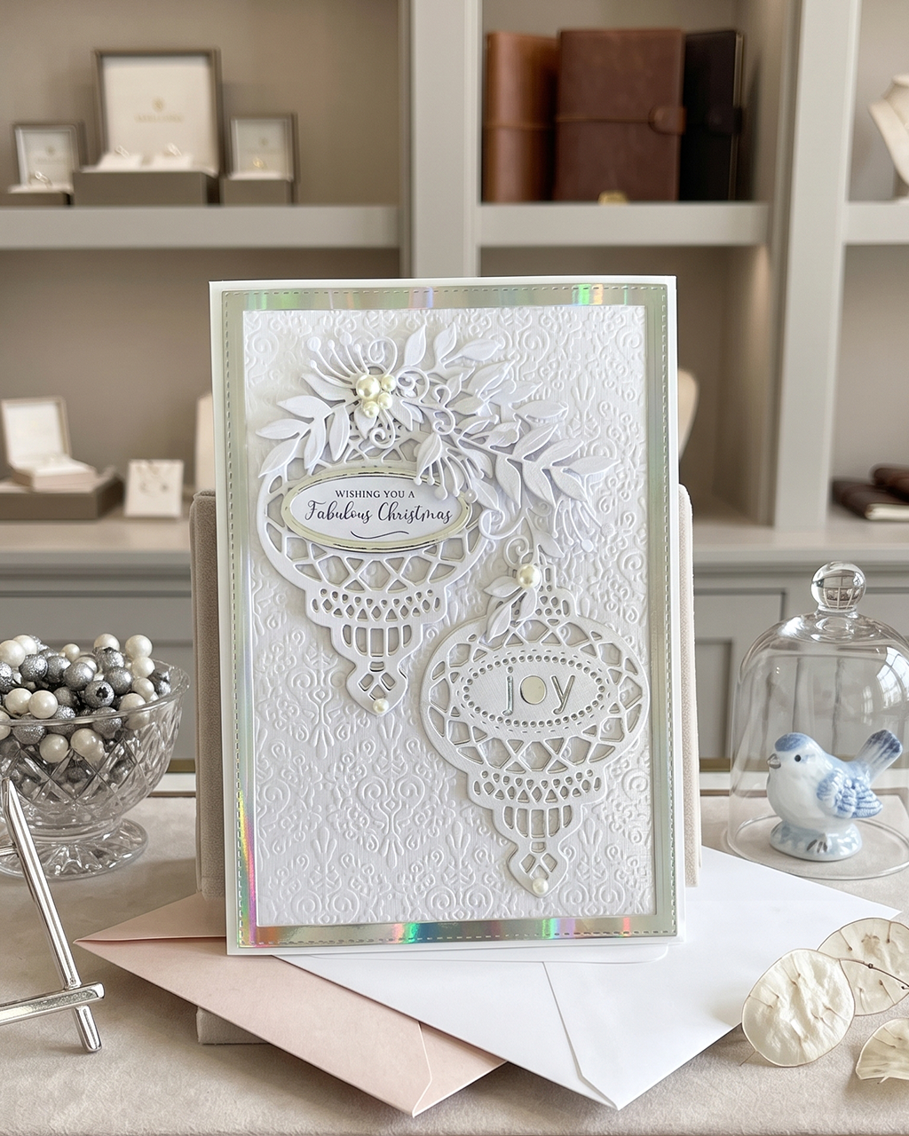 A handmade white Christmas greeting card with intricate laser-cut ornament and floral designs, displayed on a tabletop with envelopes and holiday decor in the background. Made in NZ by Del Cards NZ.