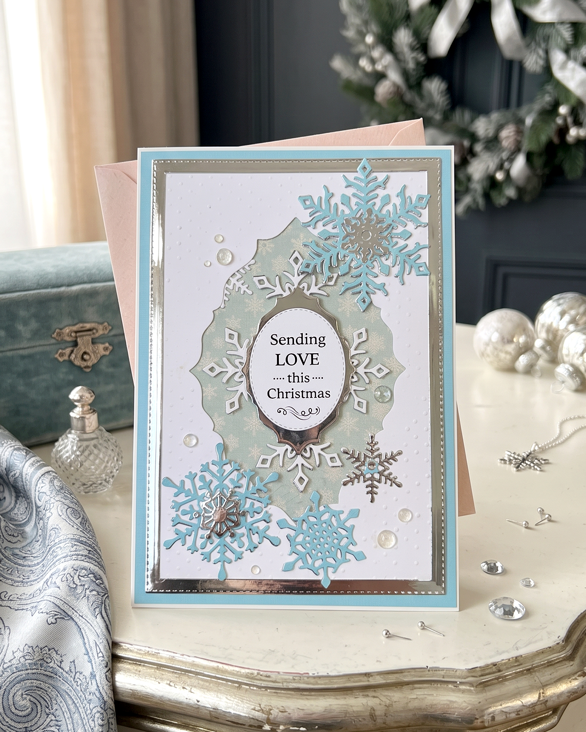 A handmade Christmas greeting card with layered light blue and silver snowflakes, a pale green watercolor wash, and a central oval reading 'Sending LOVE this Christmas' by Del Cards NZ.