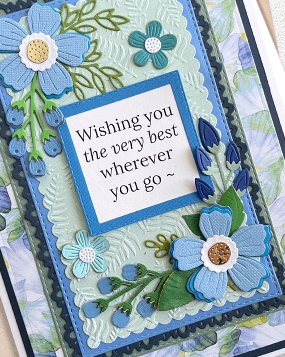A close-up of a handmade layered card with embossed floral details and a central white square that reads, 'Wishing you the very best wherever you go ~'. The card uses light blue, teal, navy, green, and white, with a scalloped blue border.