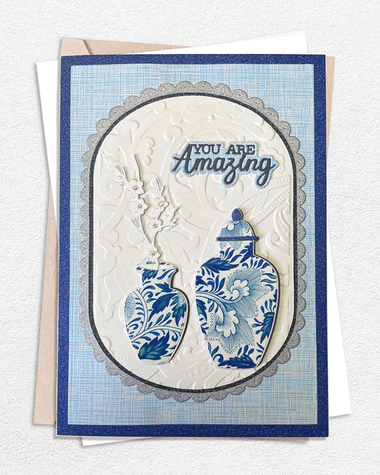 Handmade luxury greeting card in classic navy blue and ivory, featuring a textured cross-hatch navy cardstock base with a silver-edged scalloped oval frame at the centre. The raised ivory oval showcases hand-embossed cream paper and two detailed blue and white chinoiserie ginger jar illustrations. A delicate dried or paper branch spray adds height. Sentiment reads "YOU ARE Amazing" in bold and script lettering. White and kraft envelopes visible beneath. Unique handmade card made in New Zealand.
